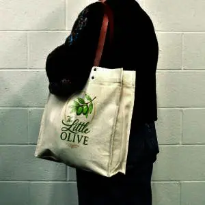 person in black jumper carrying natural colour tote bag with brown leather handles and logo print on panel of the bag