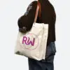 person in black jumper and blue jeans holding natural coloured bag with leather handles on shoulder. Bag has metal rivets