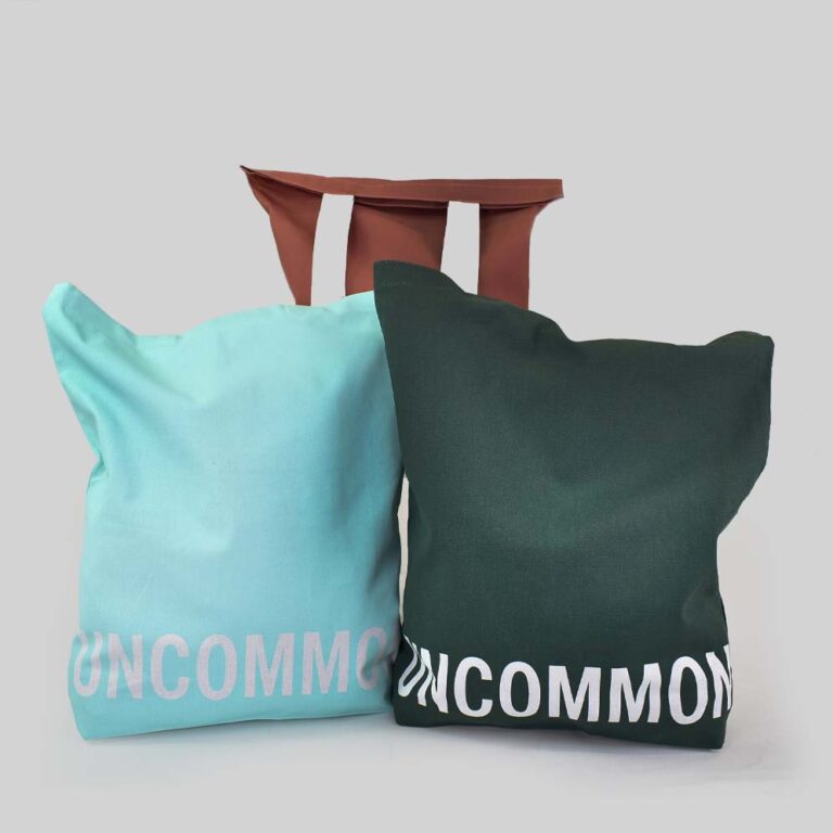 Three bags shown one in turquoise, one in rust red and the other in dark green both have white handles and white text printed along the bottom panel of the bag that says UNCOMMON