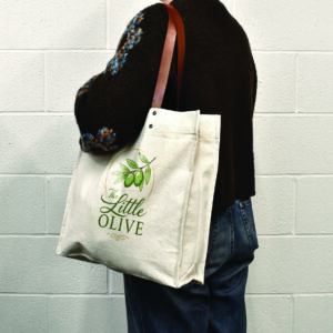 person in black jumper carrying natural colour tote bag with brown leather handles and logo print on panel of the bag