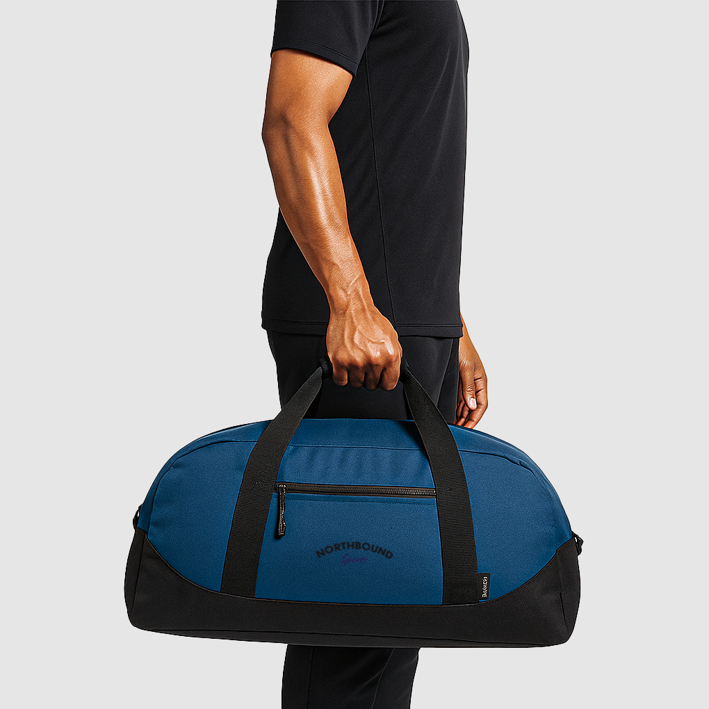 side view of person wearing a black t shirt holding a blue sports bag with black base and short carry handle. Side zipped pocket with logo print
