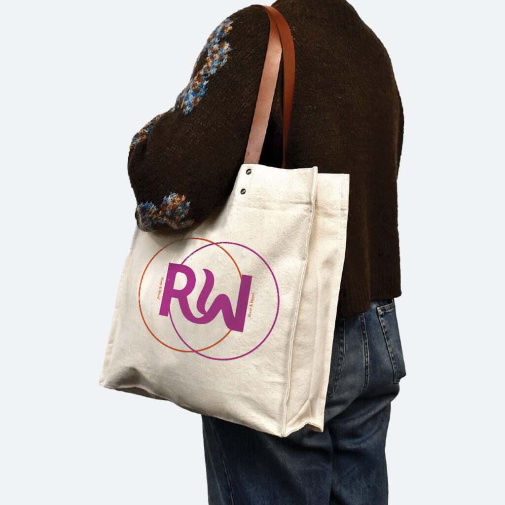 person in black jumper and blue jeans holding natural coloured bag with leather handles on shoulder. Bag has metal rivets