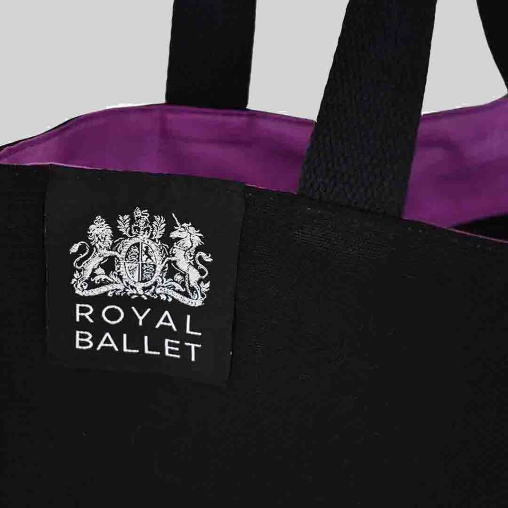 Woven label shown on top hem of black tote bag with inside purple lining. Woven label is silver Royal Ballet logo