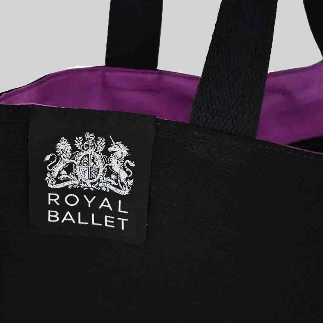 Woven label shown on top hem of black tote bag with inside purple lining. Woven label is silver Royal Ballet logo